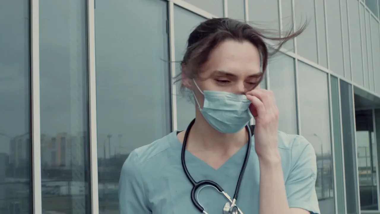 A nurse in scrubs walking out of a hospital, taking the mask down