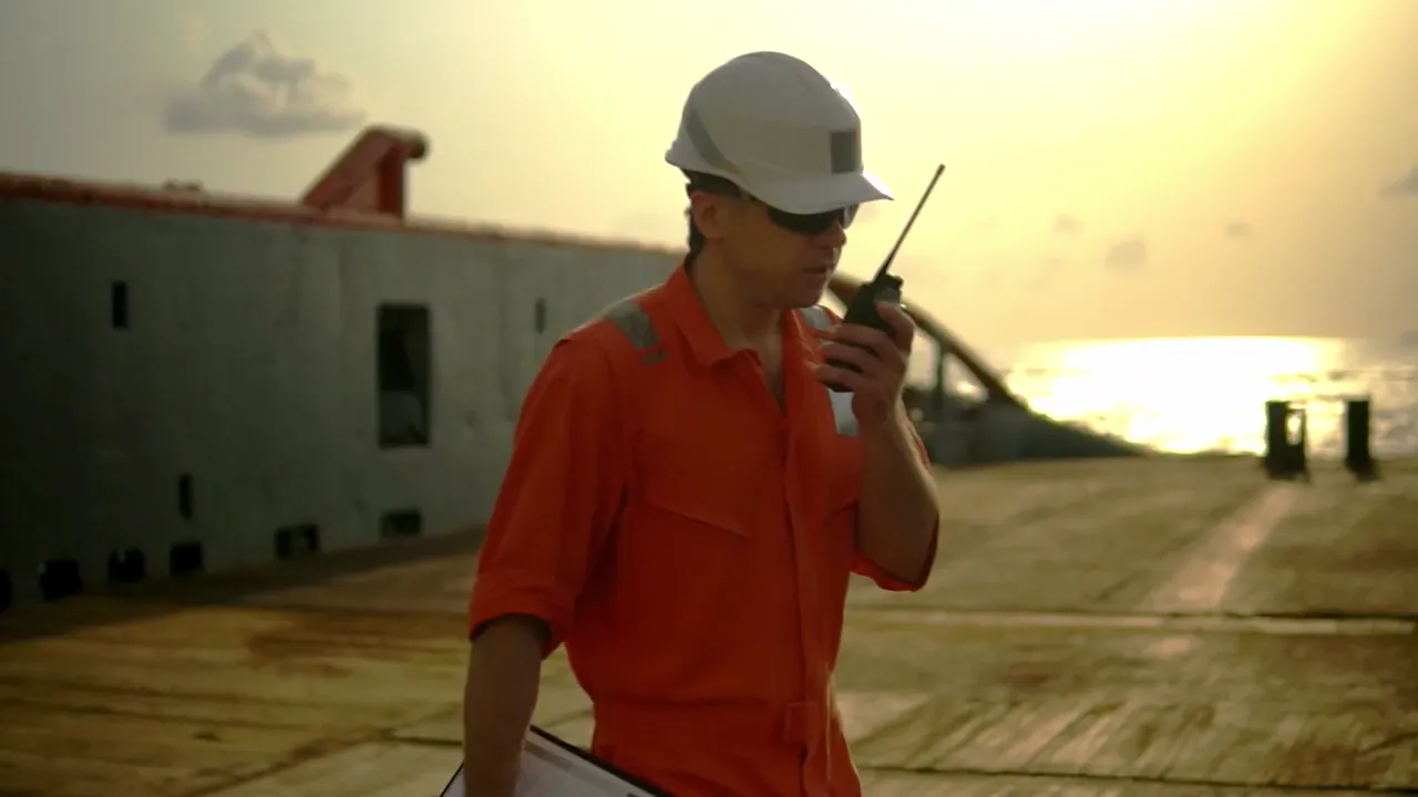 A worker in high-visibility coveralls and a hard hat on a deck at sunset, calling in on a radio